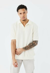 Polo shirt crème à manches courtes avec un tissu texturé et un col classique. Présente une coupe décontractée et un détail de logo subtil près du col.