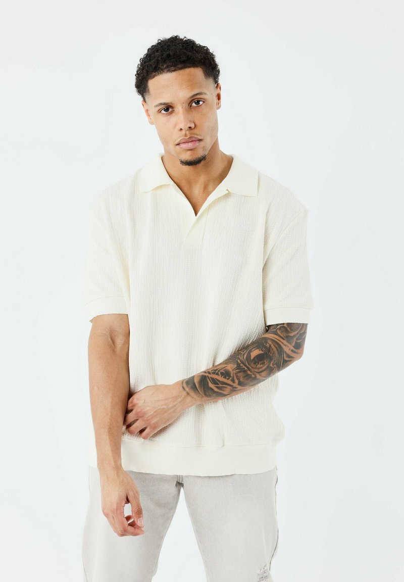 Polo shirt crème à manches courtes avec un tissu texturé et un col classique. Présente une coupe décontractée et un détail de logo subtil près du col.