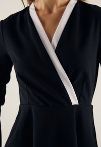 Abito nero con un trim bianco a forma di V sul collo, vita aderente e maniche lunghe, mostrato dalla collo alla vita su una persona.