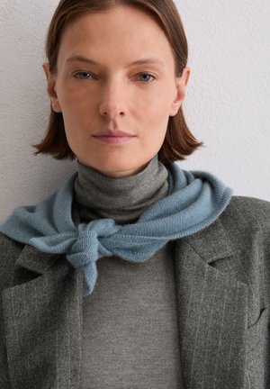 Szary wełniany blezer na szarym golfie, z jasno niebieskim szalikiem w sploty ułożonym wokół szyi, na tle teksturowanej białej ściany.