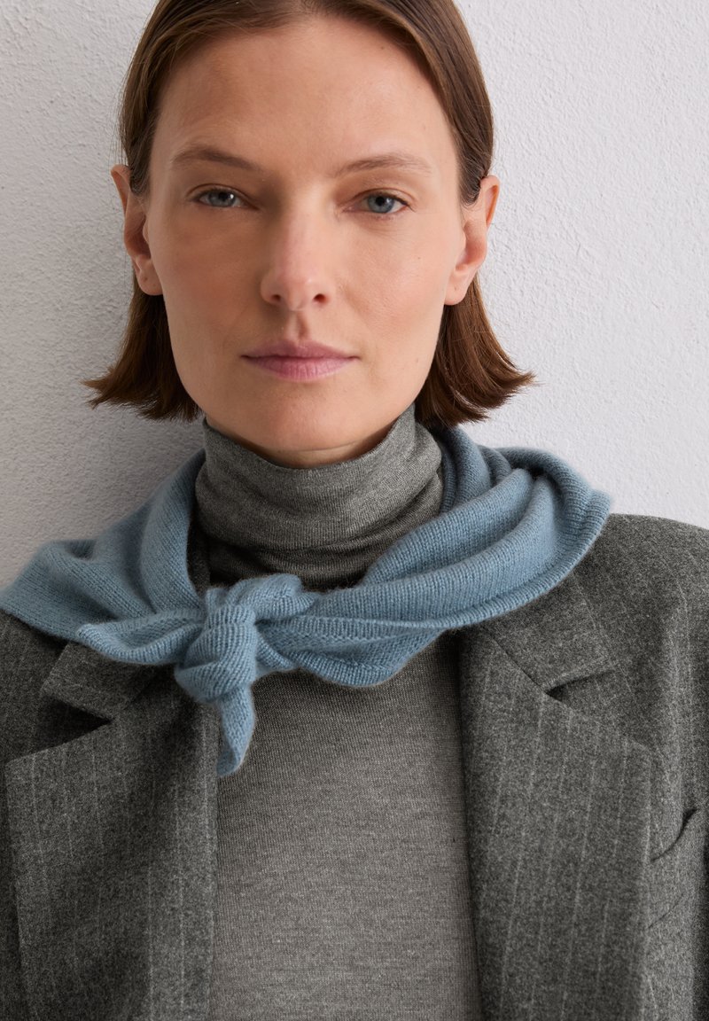 Szary wełniany blezer na szarym golfie, z jasno niebieskim szalikiem w sploty ułożonym wokół szyi, na tle teksturowanej białej ściany.
