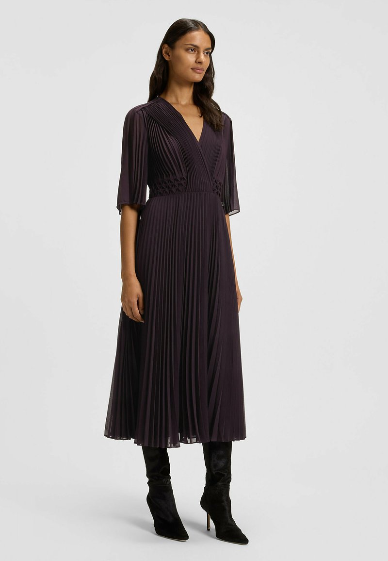 Syvän liilanvärinen plissé-mekko, jossa on V-kaula-aukko, lyhyet hihat ja teksturoitu vyötärön yksityiskohta. Yhdistetty tyylikkäisiin mokkanahkaisisiin korkokenkiin.