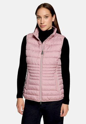 Femme portant une veste matelassée sans manches rose clair sur une chemise noire à manches longues, debout devant un fond clair uni.