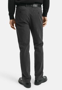 Donkergrijze broek van textuurstof, met twee achterzakken met knopen en een recht model. Draagt zwarte schoenen.