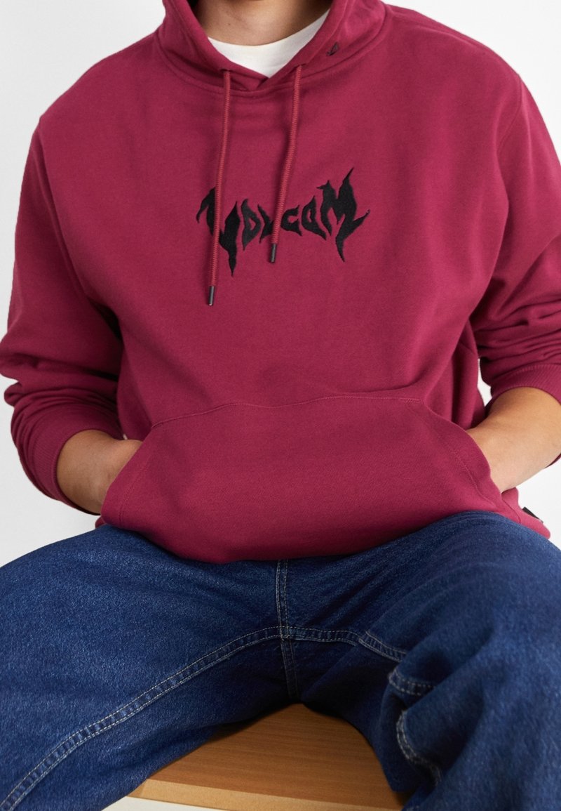 Felpa maroon con logo grafico nero, cappuccio con cordino, tasca a canguro e polsini a coste. Indossata con jeans blu.