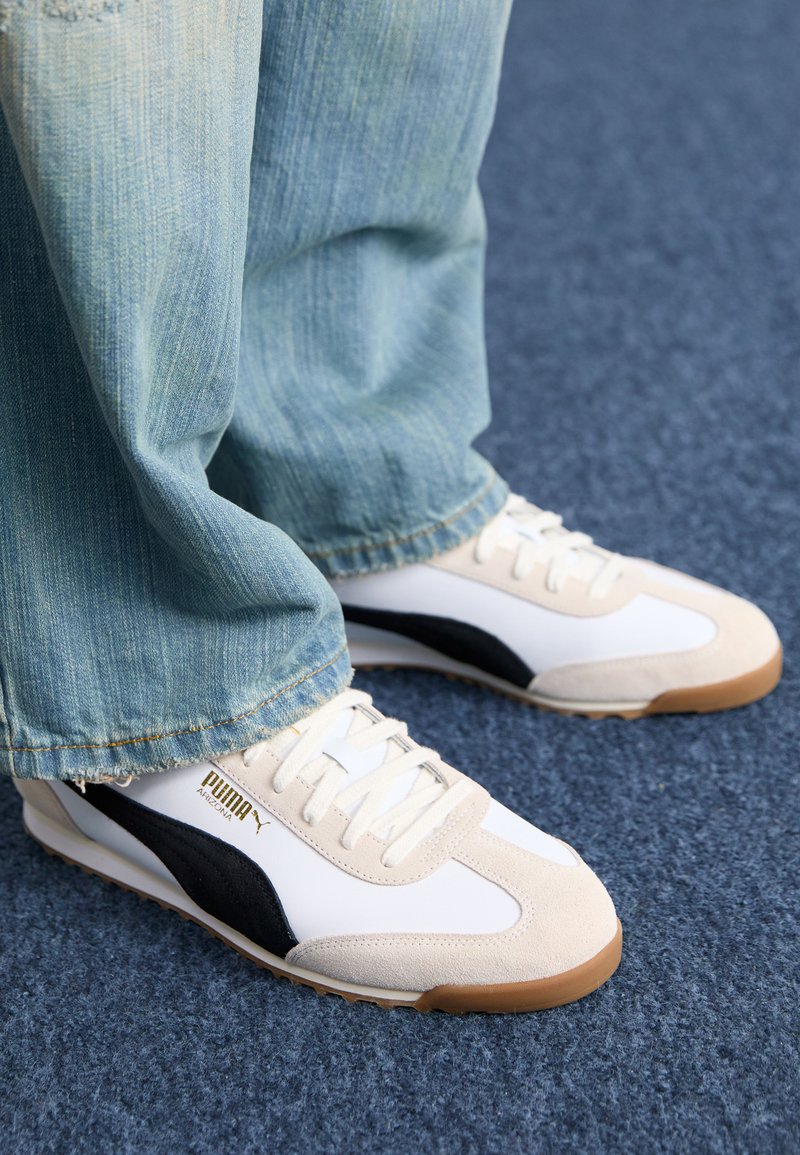 Baskets blanches et beiges avec des détails de rayures noires, comportant un panneau en suède texturé, une semelle en caoutchouc et des lacets blancs, assorties à un denim bleu.