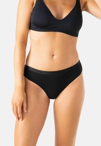 Femme portant un soutien-gorge noir sans coutures et une culotte de style bikini assortie sur un fond uni.