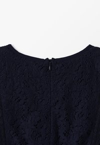 Námořnická modrá krajková šaty s kulatým výstřihem a skrytým zipem na zádech. Má květinové vzory a strukturovanou látku pro vizuální zajímavost.