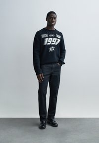 Maglione blu navy con testo grafico bianco e design a stella, abbinato a jeans scuri e scarpe nere. Texture liscia, vestibilità casual, posa in piedi.