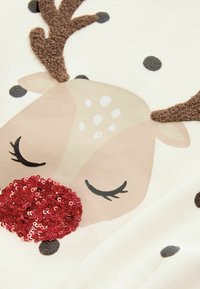 Visage de renne endormi sur tissu avec des bois marron texturés, yeux fermés, taches blanches, points noirs et nez rouge pailleté.