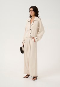 Cremfarbener, kurz geschnittener Blazer mit großen Knöpfen, kombiniert mit drapierten, weitgeschnittenen Hosen. Schwarze Riemchensandalen und eine strukturierte Schwarz-Weiß-Clutch.