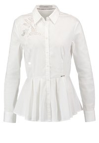 Chemise blanche à manches longues avec un corsage ajusté et un ourlet à basque plissé. Comprend une fermeture à boutons et un détail en dentelle florale sur le côté gauche.