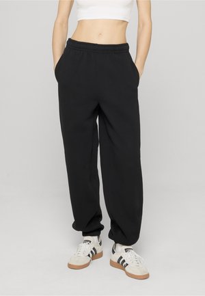 Persoon draagt zwarte losse joggingbroek, wit cropped top en witte sneakers met zwarte strepen, staand met handen in de zakken.