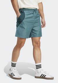 Tyrkysové šortky s elastickým pásom a šnúrkou, s temnomodrou bočnou kapsou. V kombinácii s bielymi teniskami a čierno pruhovanými ponožkami.