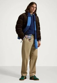 Νεαρός άνδρας φορώντας ένα μαύρο μπουφάν, μπλε πλεκτό πουλόβερ, καρό πουκάμισο, ανοιχτόχρωμο παντελόνι, μπλε ριγέ κασκόλ και πράσινα αθλητικά παπούτσια, στέκεται.