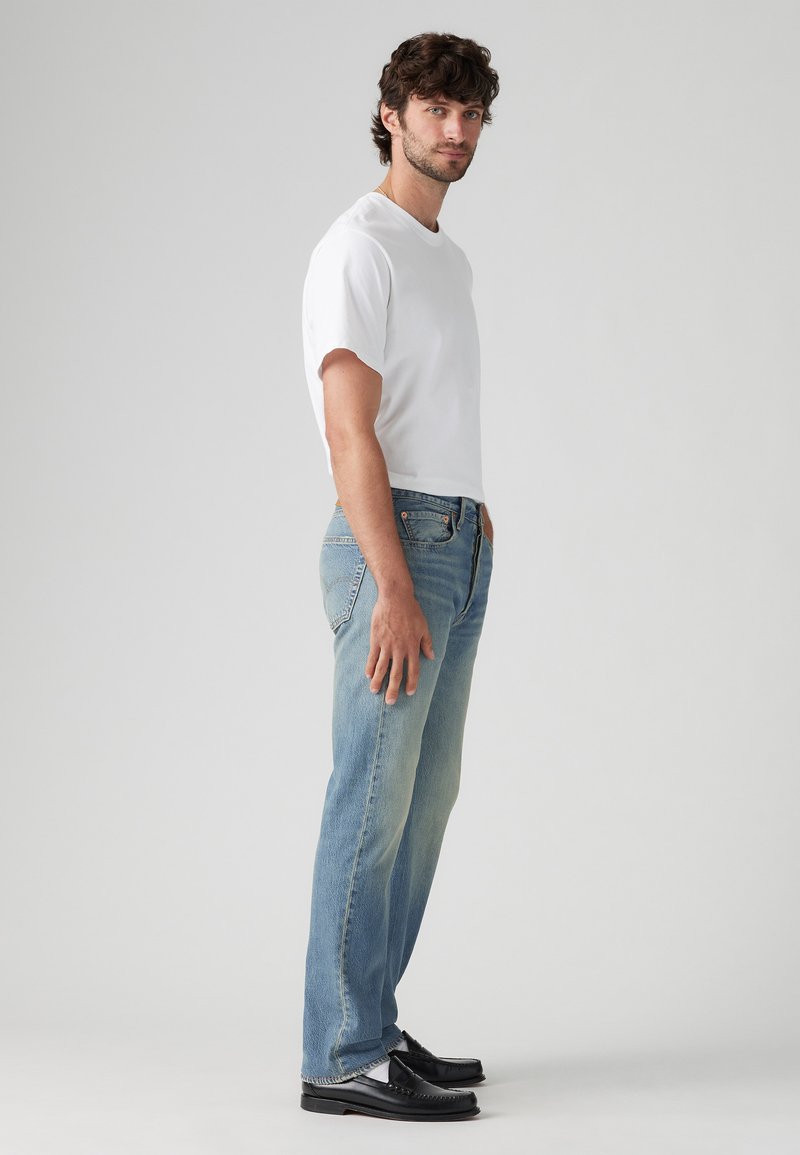 T-shirt blanc à col rond, jean en denim bleu clair légèrement usé, coupe droite, porté avec des mocassins en cuir noirs, vue de profil contre un fond uni.