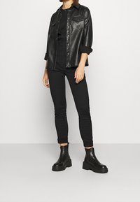 Czarna koszula kurtka z faux skóry z dwiema kieszeniami na piersi, w połączeniu z czarnymi dopasowanymi spodniami i masywnymi czarnymi botkami za kostkę.