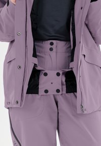 Set di abbigliamento esterno di colore viola chiaro, composto da una giacca con zip e chiusure a scatto, foderata di nero, e pantaloni abbinati con bottoni e cintura elastica.