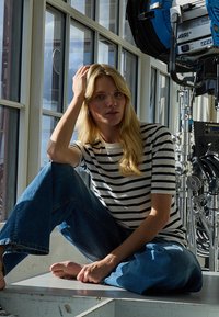 Femme blonde en chemise rayée et jean, assise pieds nus sur une plateforme près d'une grande fenêtre, avec un équipement d'éclairage de cinéma à proximité.