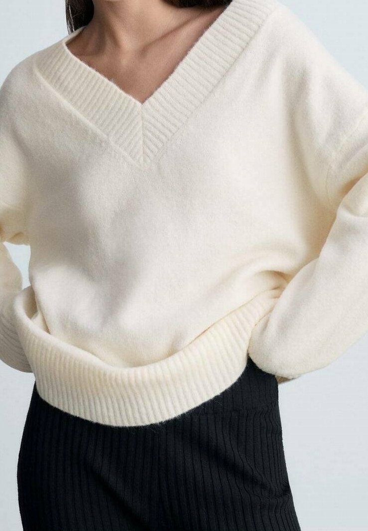 Pull crème, doux, avec un décolleté en V profond et un ourlet côtelé. Présente un tricot texturé et une coupe décontractée, associé à un pantalon noir côtelé.