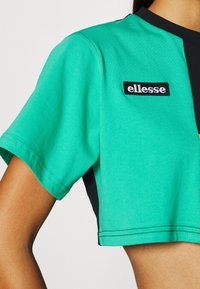 Camiseta corta verde con cuello negro y paneles laterales, que presenta un parche del logo "ellesse" en blanco en el pecho.