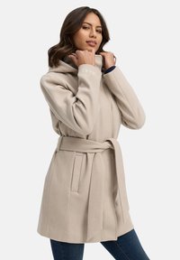 Cappotto beige con cappuccio e vita con cintura, caratterizzato da una texture morbida, due tasche laterali e dettagli di cucitura verticale. Logo ricamato sul cappuccio.
