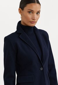 Mornarsko modra blazer jakna v strukturirani zasnovi, z ovratnikom s križnimi robovi, enim gumbom za zapiranje in sprednjimi žepi z zavihkom ter gladko teksturo.