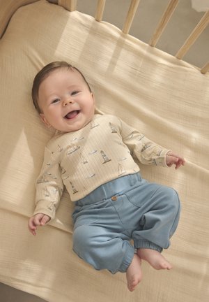 Light beige long-sleeve shirt with sailboat pattern, paired with soft blue pants. Baby smiles on a textured beige blanket in a crib.