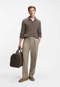 Bruine gebreide polo, beige wijde broek en bruine sneakers; houdt een donkerbruine sporttas met leren accenten vast. Textuurdetails van de stof zichtbaar.
