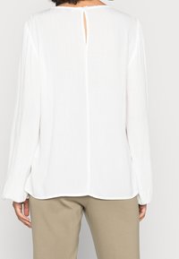 Blusa bianca a maniche lunghe con tessuto strutturato, chiusura posteriore a occhiello e vestibilità rilassata, abbinata a pantaloni beige.