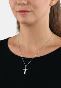 Silverfärgad korshalsband med en texturerad design på en tunn kedja, bärs över en svart ribbad topp. Anmärkningsvärd glans och minimalistisk form.