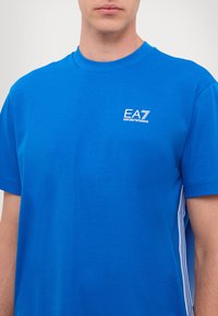 Modrý bavlněný tričko s krátkým rukávem. Na levé části hrudi je bílé logo EA7 a po stranách jsou vertikální bílé pruhy.