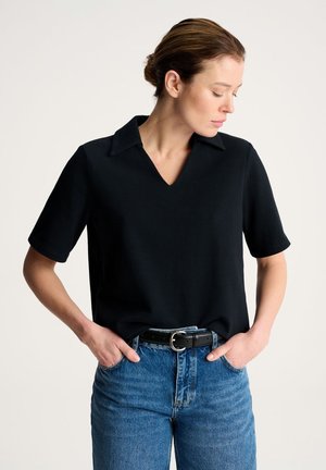 Kobieta z rękami w kieszeniach, ubrana w czarną koszulę z krótkim rękawem i kołnierzykiem, włożoną do niebieskich dżinsów z czarnym paskiem, patrząca w dół na bok.