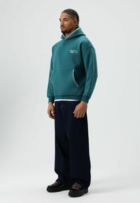 Groenblauwe capuchontrui met een zak aan de voorkant, met contrasterende stiksels en een losse pasvorm, gecombineerd met losse donkere spijkerbroek en zwarte schoenen.