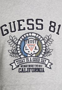 Sudadera gris con el texto "GUESS" en negrita, logo bordado con acentos en azul, verde y blanco, y diseños florales. El texto dice "Hoy es un buen día."