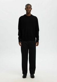 Černý svetr s kulatým výstřihem, dlouhými rukávy a žebrovanými manžetami, doplněný volnými černými kalhotami a černými botami.