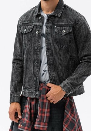 Mann trägt schwarze ausgebleichte Jeansjacke über einem Batikshirt, hält ein rot-schwarz kariertes Hemd um die Taille.