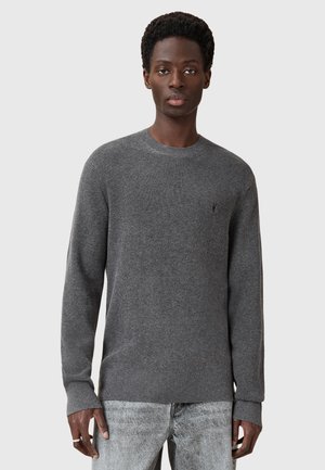 Grauer, strukturierter Strickpullover mit Rundhalsausschnitt und langen Ärmeln. Mit einem kleinen Logo auf der Brust. Kombiniert mit hellgrauen Jeans.