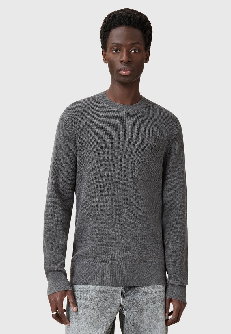 Szary, strukturalny sweter z dzianiny o okrągłym dekolcie i długich rękawach. Posiada małe logo na klatce piersiowej. Połączony z jasnoszarymi dżinsami.