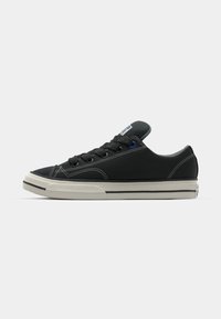 Black canvas sneaker with white rubber sole, round toe, and contrasting stitching. Features black laces and a blue accent on the tongue.