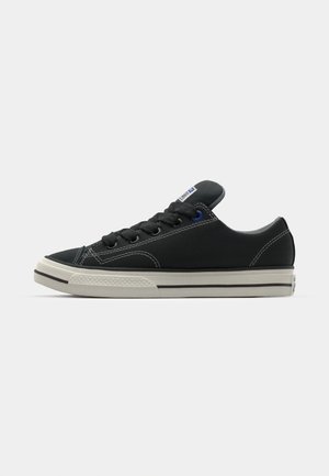 Sneaker nera in tela con suola in gomma bianca, punta tonda e cuciture in contrasto. Presenta lacci neri e un accento blu sulla linguetta.