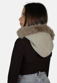 Sciarpa con cappuccio in maglia beige con rifinitura in morbida pelliccia sintetica. Presenta un design testurizzato ed è indossata sopra una maglia nera, mettendo in risalto la sua forma.