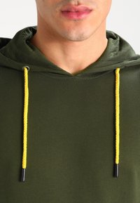 Volto inferiore di una persona che indossa un felpa color verde oliva con cordini gialli luminosi su uno sfondo chiaro e semplice.