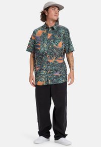 Camisa de botones de manga corta en verde oscuro con un vibrante estampado tropical, que presenta hojas de palma y patrones de atardecer; combinada con pantalones negros.