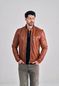 Brun læderjakke med høj krave, lynlåslommer og tætsiddende design. Båret over en mørk sweater og grå bukser. Glat tekstur.