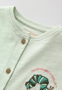 T-shirt bébé vert clair avec boutons en bois et personnage brodé de "La très affamée chenille" sur la poitrine.