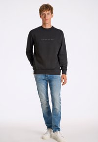 Felpa nera con colletto e polsini a coste, caratterizzata da un logo grigio discreto. Abbinata a jeans azzurri chiari e sneakers bianche.