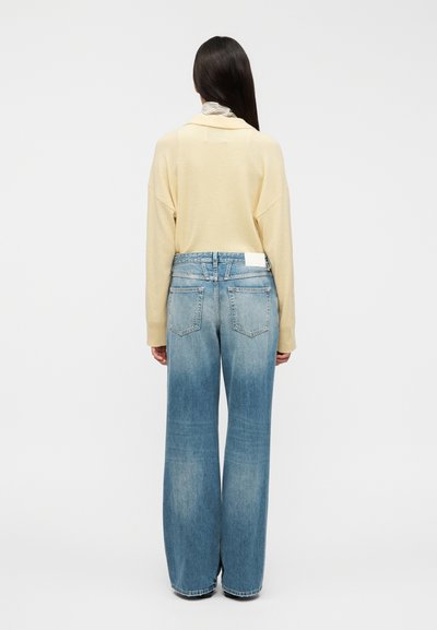 Jersey grande de color amarillo claro, vaqueros holgados azul claro. Piernas anchas y denim descolorido destacados. Diseño sencillo, sin logotipos ni patrones visibles.