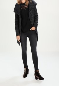 Abrigo de lana gris oscuro con cuello oversized, blusa de encaje negra, jeans ajustados negros y botines de ante negros con detalle de cremallera dorada.
