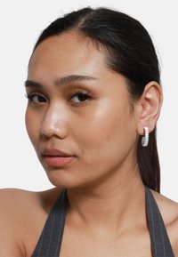 Boucles d'oreilles en argent en forme de créole avec un design texturé, présentant une légère courbure et une finition polie, portées par une personne sur fond blanc.